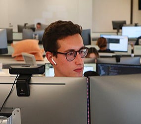 A man with glasses and earbuds in working behind his computer in an office
